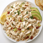 Creamy homemade chicken salad served on a white plate with celery, grapes, pecans, and crackers, garnished with fresh dill and a lemon wedge.